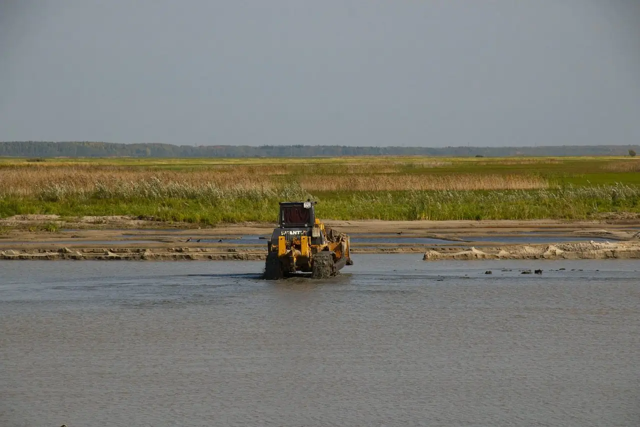 Bulldozer working at the edge of a body of water beside tall grass