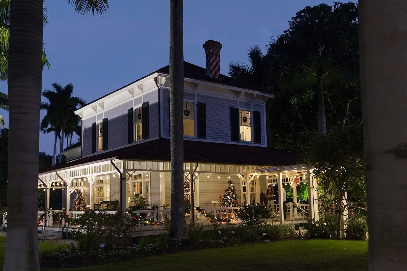 White two-story house with a wide porch lit at dusk among palms